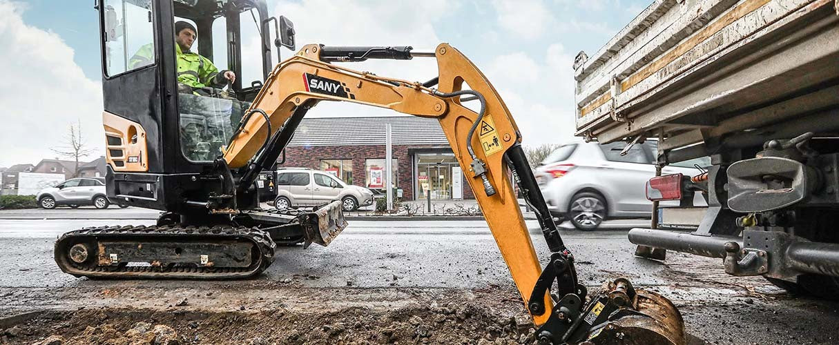 Operator minikoparki wykonuje wykop przy krawędzi jezdni obok zaparkowanej ciężarówki.