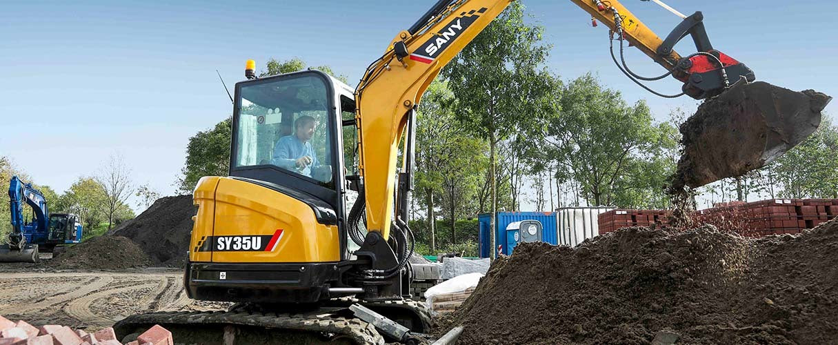 Minikoparka SANY przenosi ziemię na placu budowy otoczonym drzewami i materiałami.