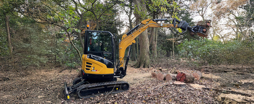 Mała koparka gąsienicowa w lesie podnosi pniak drzewa za pomocą chwytaka hydraulicznego.