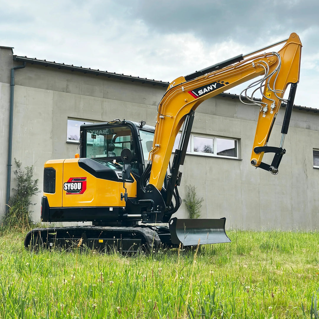 Minikoparka SANY SY60U na gąsienicach, ujęcie boczne z widocznym ramieniem hydraulicznym i lemieszem, maszyna zaparkowana przy budynku na trawie.