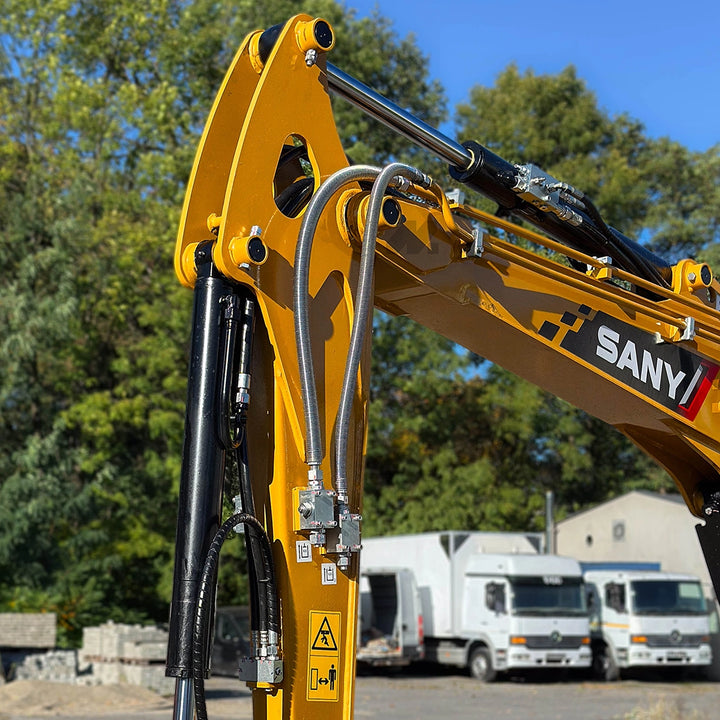 Wysięgnika minikoparki Sany SY35U – zbliżenie na siłownik hydrauliczny i przewody w stalowym oplocie na tle zielonych drzew.